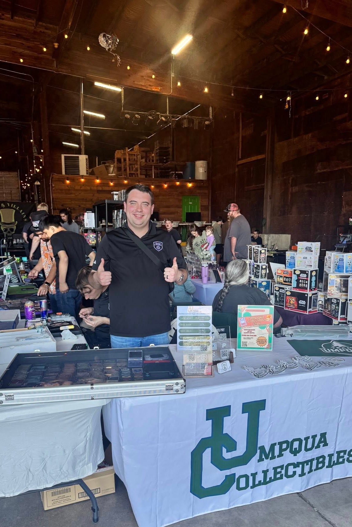 A man wearing a black Umpqua Collectibles polo shirt stands behind a white table draped with a matching Umpqua Collectibles tablecloth at a booth inside a rustic barn.