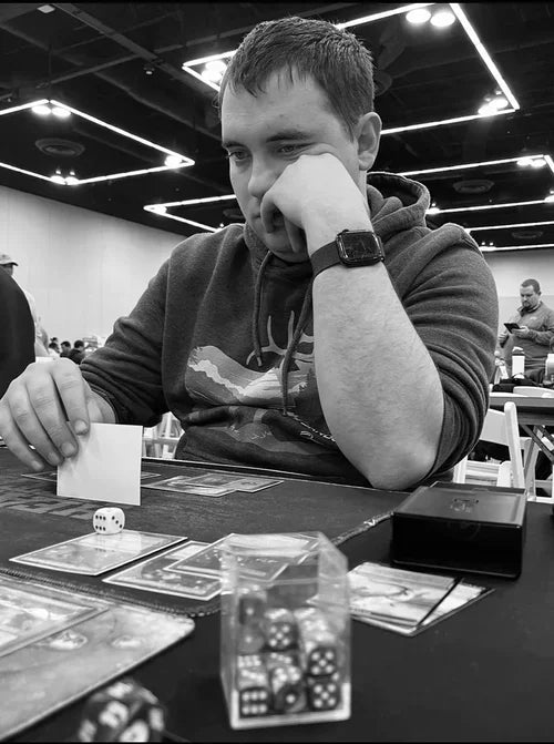 A man in a dark hoodie sits at a table with cards and dice, holding a small white card in a black-and-white event setting.